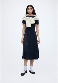Femme portant une robe midi noire à manches courtes, une écharpe blanche sur les épaules, des chaussettes blanches et des chaussures Mary Jane noires, debout devant un fond uni.