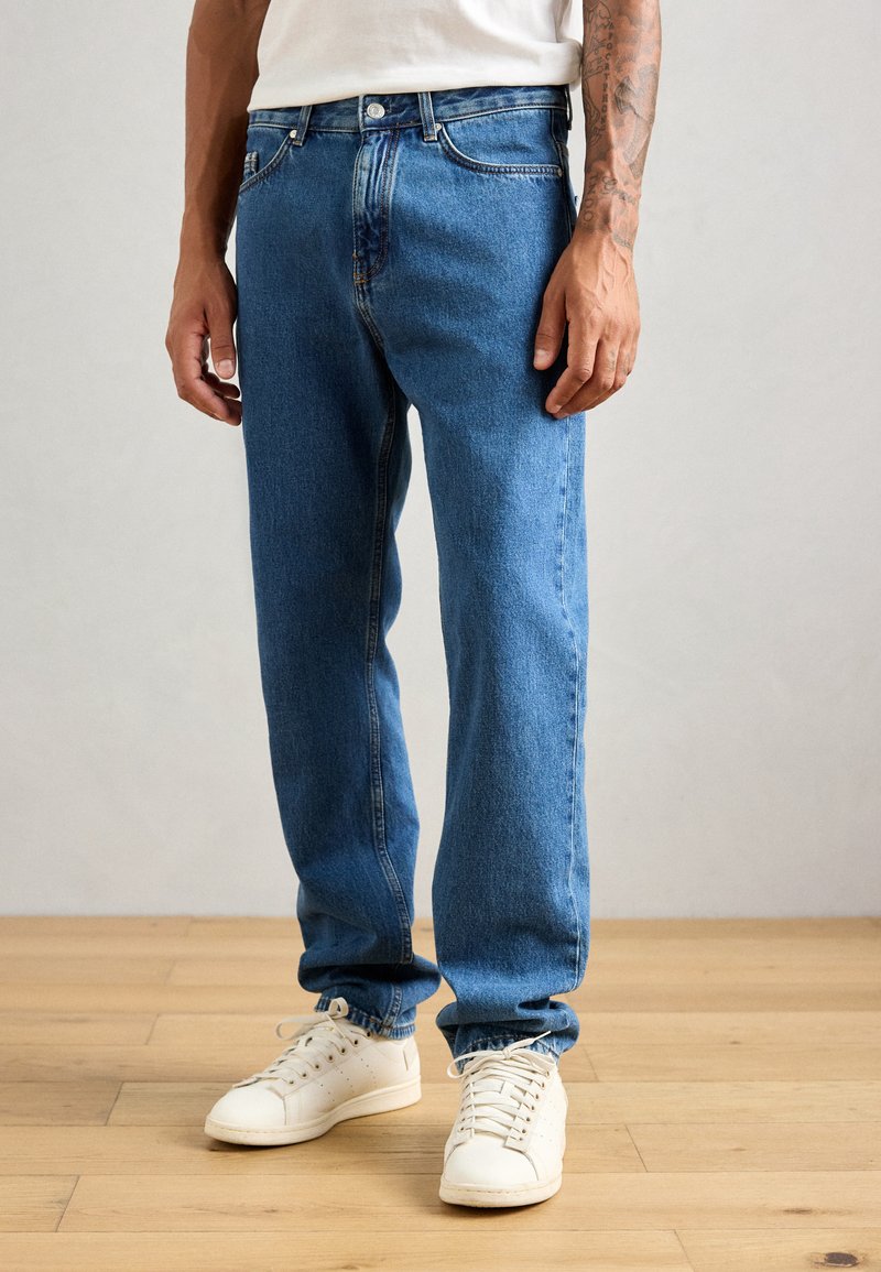 Mann i rette jeans i blått og hvite joggesko som står på tregulv mot en enkel vegg.