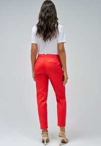 Mujer con cabello largo y oscuro, vestida con una camiseta blanca, pantalones rojos y sandalias de tacón beige, de pie mirando hacia atrás sobre un fondo liso.