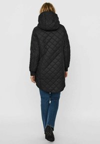 Manteau long matelassé noir avec capuche, arborant un motif en losanges, des poignets élastiques et un ourlet arrondi. Associé à un jean bleu et des bottes noires.