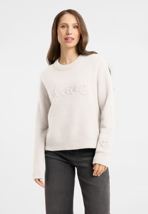 Woman wearing a white sweater with "love" text and a heart design on the sleeve, paired with dark gray pants, standing against a white background.