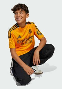Maillot de soccer orange vif avec des rayures noires et un logo, associé à un pantalon noir et des baskets blanches. Le modèle est à genoux, souriant, sur un fond neutre.