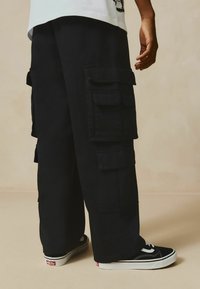 Des pantalons cargo noirs présentent de grandes poches multiples, une coupe décontractée et sont associés à des baskets noires avec une semelle blanche et des détails de couture.