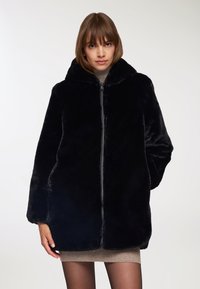 Cappotto di pelliccia nera con cappuccio, dotato di zip frontale, maniche ampia e una texture morbida e pelosa. Indossato sopra un vestito beige.