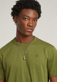 T-shirt verde oliva con collo a giro, texture a costine sul colletto, piccolo logo sul petto e collana con pendente in argento.