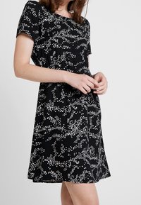 Svart klänning med vitt blommönster; korta ärmar, rund halsringning, utsvängd silhuett, mjukt tyg, knälång.