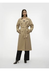Beige trenchcoat med dobbeltknapper, et knyttet belte i livet og store lommer, kombinert med svarte vide bukser og hælsko.