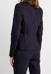 Blazer blu navy con design strutturato, caratterizzato da una silhouette aderente e dettagli delle cuciture sottili. La vista posteriore mostra una trama di tessuto liscia e linee pulite.