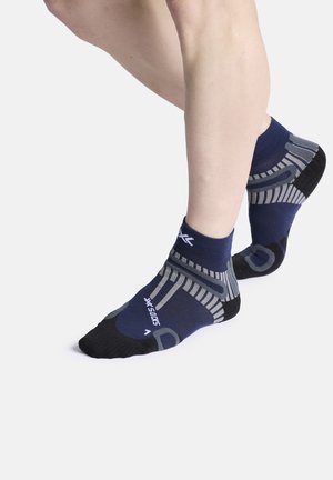 Personne portant des chaussettes de sport basses bleu marine et grises avec des orteils et un talon noirs, debout sur la pointe des pieds sur fond blanc.