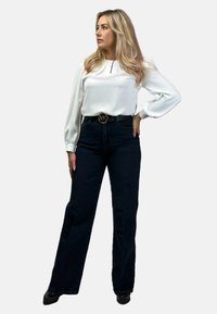 Biała bluzka o długich rękawach z dekoltem w kształcie łezki, w połączeniu z ciemnoniebieskimi spodniami z wysokim stanem i czarnym paskiem z okrągłym logo.