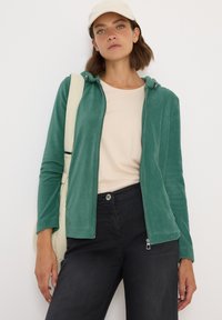 Veste zippée en velours côtelé vert avec capuche, associée à un t-shirt beige et un pantalon noir ; portant une casquette crème et tenant un sac.