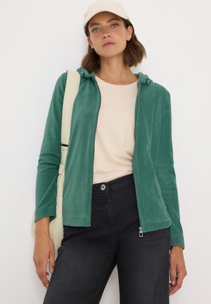 Veste zippée en velours côtelé vert avec capuche, associée à un t-shirt beige et un pantalon noir ; portant une casquette crème et tenant un sac.