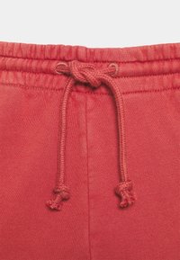 Röda bomullssweatpants med dragsko i midjan. Tyget är mjukt med en slät yta. Dragskoändarna är knutna med små tofsar.