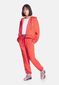 Jasnopomarańczowa bluza z kapturem i dopasowane spodnie dresowe wykonane z miękkiego materiału, z przednim zamkiem błyskawicznym i sznurkami do regulacji. Noszone z różowymi butami sportowymi.