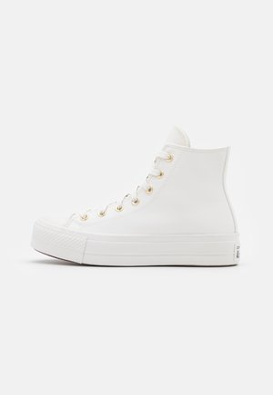Baskets montantes blanches avec œillets dorés, semelle épaisse en caoutchouc et lacets blancs, vues de profil sur un fond uni.