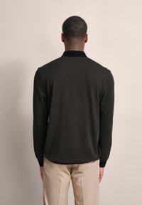 Dunkelgrüner Strickpullover mit einem marineblauen Kragen, gerippten Bündchen und einer strukturierten Oberfläche. Getragen mit hellbeigen Hosen, die ein schlichtes Design aufweisen.