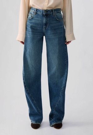 Personne portant un jean bleu à jambes larges et une chemise à manches longues crème, debout face à un fond clair uni.