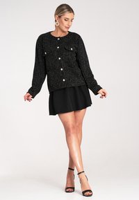 Czarna, teksturowana kurtka z wzorem w kratkę, wyposażona w srebrne guziki i dwie przednie kieszenie. W połączeniu z czarną dopasowaną spódnicą.