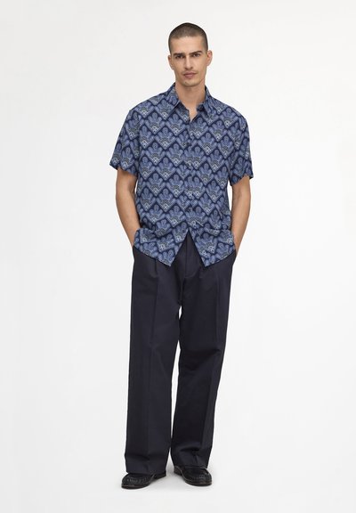 Homme aux cheveux courts portant une chemise bleue à motifs à manches courtes et un pantalon ample bleu marine foncé, debout les mains dans les poches.