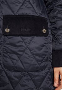 Marineblauwe gewatteerde jas met een corduroy zak met twee knopen, merkopdruk en geribde boorden voor extra textuur en detail.