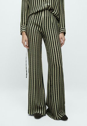 Femme portant un pantalon large à rayures verticales vert olive et noir et un haut à manches longues assorti, debout devant un fond uni.