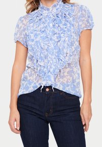Blouse bleu clair avec un motif floral, devant volé, et col haut. Manches courtes et tissu transparent complètent un jean en denim sombre.