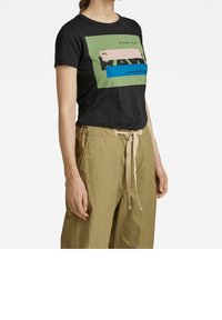 Kvinna som bär en svart grafisk t-shirt med gröna och blå block samt khaki byxor med dragsko, visad från axlarna till mitten av låren.