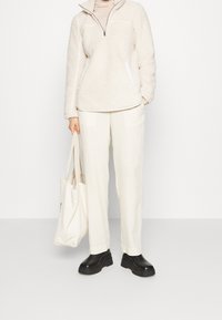 Henkilö, joka pukeutunut kermaiseen fleece-pooloon, valkoisiin housuihin ja mustiin korokepohjakenkiin, pitää valkoista kangaskassia yksiväristä vaaleaa taustaa vasten.