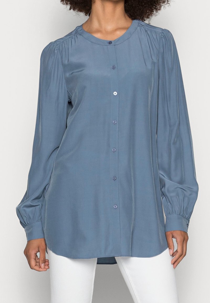 Femme portant un chemisier bleu à manches longues boutonné, avec des épaules et des poignets froncés, assorti à un pantalon blanc.