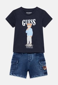 Μπλε βαμβακερό t-shirt με γραφικό αρκούδας και κείμενο "GUESS", συνδυασμένο με τζιν σορτς με πλαϊνές τσέπες και έμβλημα αρκούδας.
