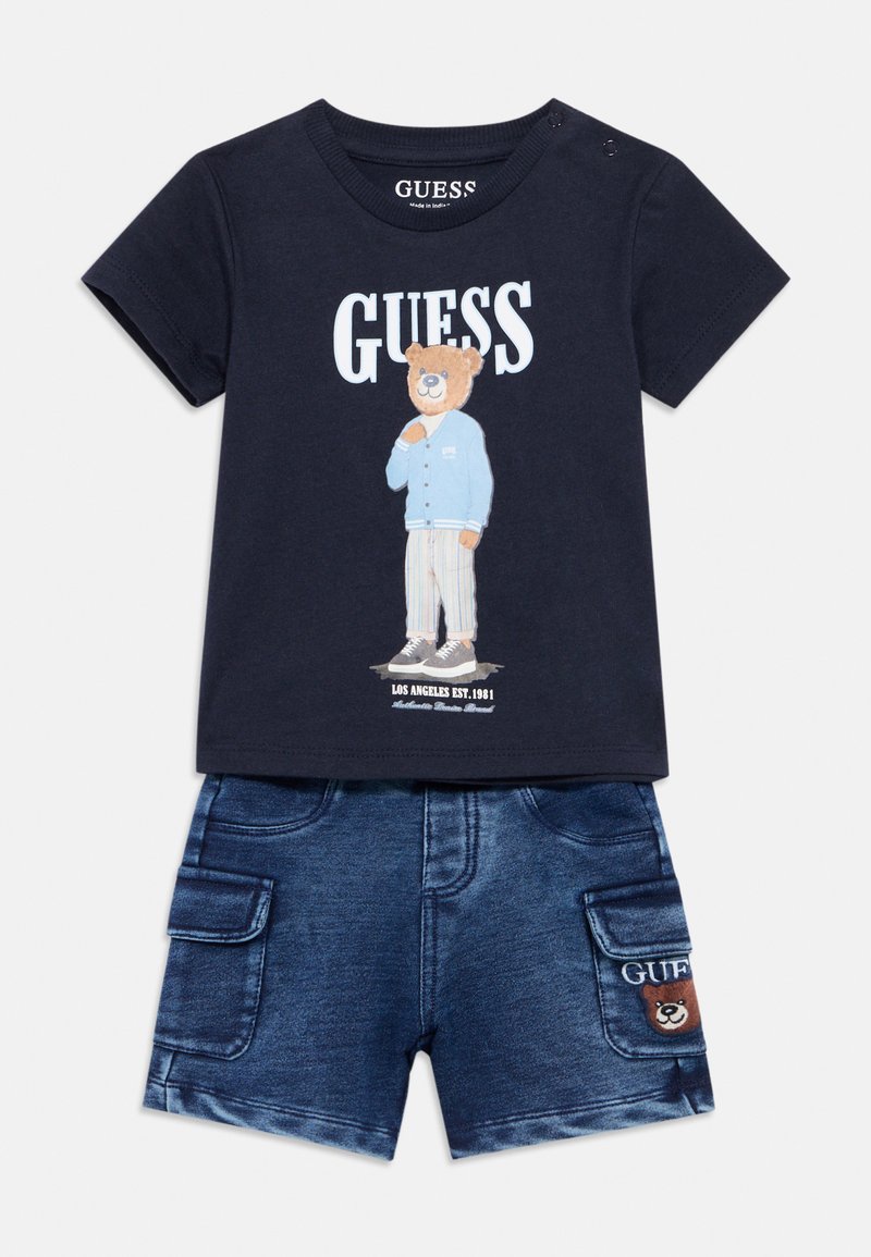 Μπλε βαμβακερό t-shirt με γραφικό αρκούδας και κείμενο "GUESS", συνδυασμένο με τζιν σορτς με πλαϊνές τσέπες και έμβλημα αρκούδας.