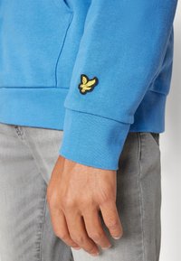 Felpa azzurro chiaro con polsini a coste e logo giallo ricamato sulla manica sinistra, abbinata a jeans grigio chiaro.