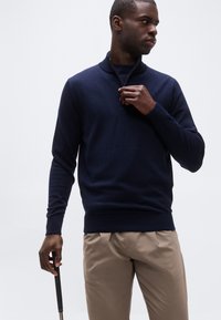 Homme ajustant la fermeture éclair d'un pull bleu marine tout en tenant un club de golf, portant un pantalon beige sur un fond clair uni.