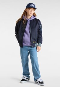 Marineblauer Bomberjacke über einem lila Hoodie, kombiniert mit hellblauen Jeans und schwarzen Sneakern. Mit Reißverschluss und Seitentaschen. Blauer Hut.