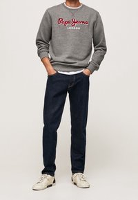 Felpa grigia con logo ricamato rosso, abbinata a jeans blu scuro e sneakers beige. Look casual e comodo con polsini a costine.
