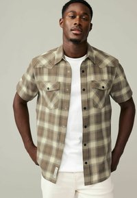 Bruin en roomkleurig geruit overhemd met korte mouwen, drukknopen, twee borstzakken en een recht gesneden zoom, gedragen over een wit t-shirt.