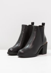 Botas de tobillo de cuero negro con paneles laterales elásticos, punta redondeada y un tacón grueso. Textura suave con un detalle dorado en la parte trasera.