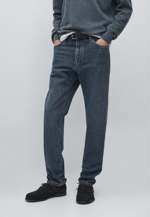 Persona con jeans blu scuro infilati in scarpe nere con i lacci, abbinati a un maglione grigio e mani in tasca su uno sfondo neutro.