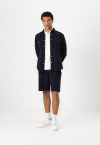 Polo alb cu mânecă scurtă sub o cămașă albastră închis, asociat cu pantaloni scurți albaștri închiși și adidași albi. Ținută simplă și casual.