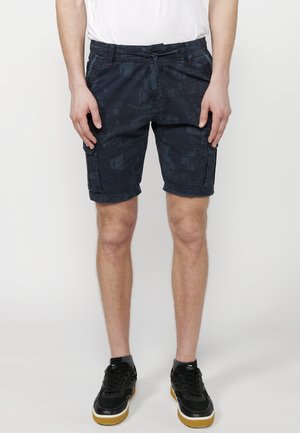 Dunkelblaue Shorts mit geometrischem Muster, leichtem Stoff, elastischem Bund und seitlichen Taschen. Tragen mit schwarzen Sneakern.