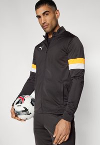 Μαύρο αθλητικό μπουφάν με πορτοκαλί και άσπρες λωρίδες στα μανίκια, κατασκευασμένο από λείο υλικό. Άντρας κρατάει μια άσπρη και μαύρη μπάλα ποδοσφαίρου.