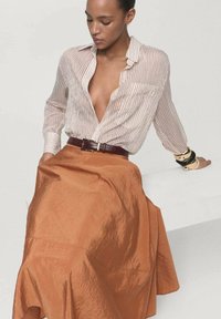 Gestreepte button-up shirt in lichtbeige met bordeauxrode strepen, gecombineerd met een glanzende oranje rok tot de knie. Bruin leren riem en armbanden.