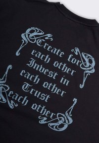 Tessuto nero con testo in stile gotico blu che recita: "Crea per l'uno e per l'altro, Investi l'uno nell'altro, Fidati l'uno dell'altro," incorniciato da decorazioni floreali agli angoli.