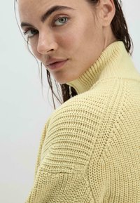 Texturiertes gelbes Strickpullover mit hohem Kragen, ausgestattet mit gerippten Akzenten und lockerer Passform, präsentiert an einem Modell mit nassem Haar.