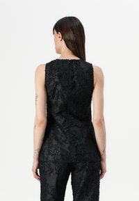 Svart ermeløs topp med et teksturert blomster mønster, rett snitt og en sentral glidelås bak. Stoffet har en glatt overflate.