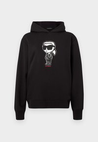 Crna kapuljača s grafikom crtića u bijeloj boji, koja nosi naočale i odijelo, s natpisom "KARL LAGERFELD" u crvenoj boji ispod.