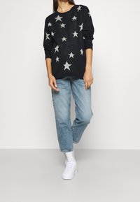 Schwarzer Strickpullover mit silbernen Sternmotiven, kombiniert mit hellblauen, locker sitzenden Jeans und weißen Turnschuhen. Der Pullover hat einen rund ausgeschnittenen Hals.