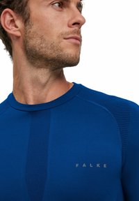 T-shirt de sport bleu avec texture côtelée, col rond et panneaux latéraux contrastants. Présente le logo FALKE en gris clair sur la poitrine.