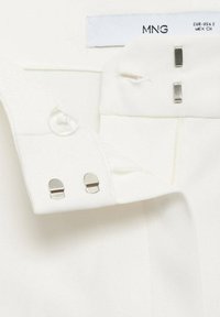 Guler de bluză albă cu un buton transparent, elemente din metal argintiu și detalii cusute. Etichetă cu "MNG" și informații despre mărime vizibile.