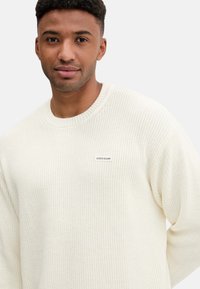 Maglione lavorato a maglia color crema con collo rotondo e trama a coste. Presenta un piccolo logo "GUESS JEANS" sul petto.